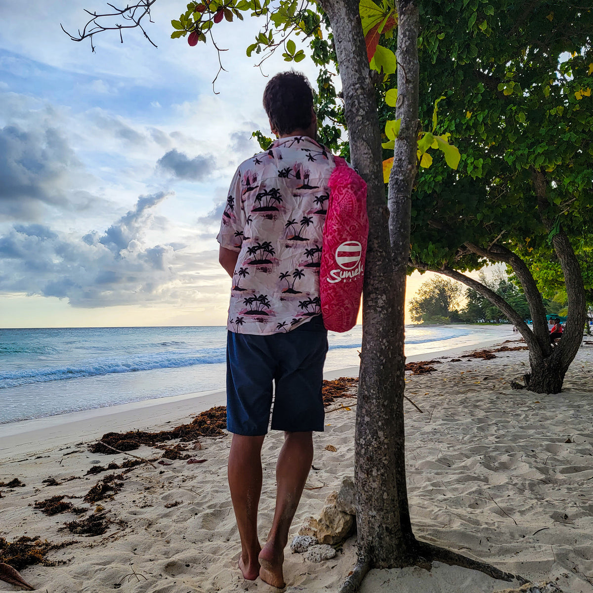 Man stands on a tropical beach carrying a pink suniela shade in it's compact carry bag over his shoulder