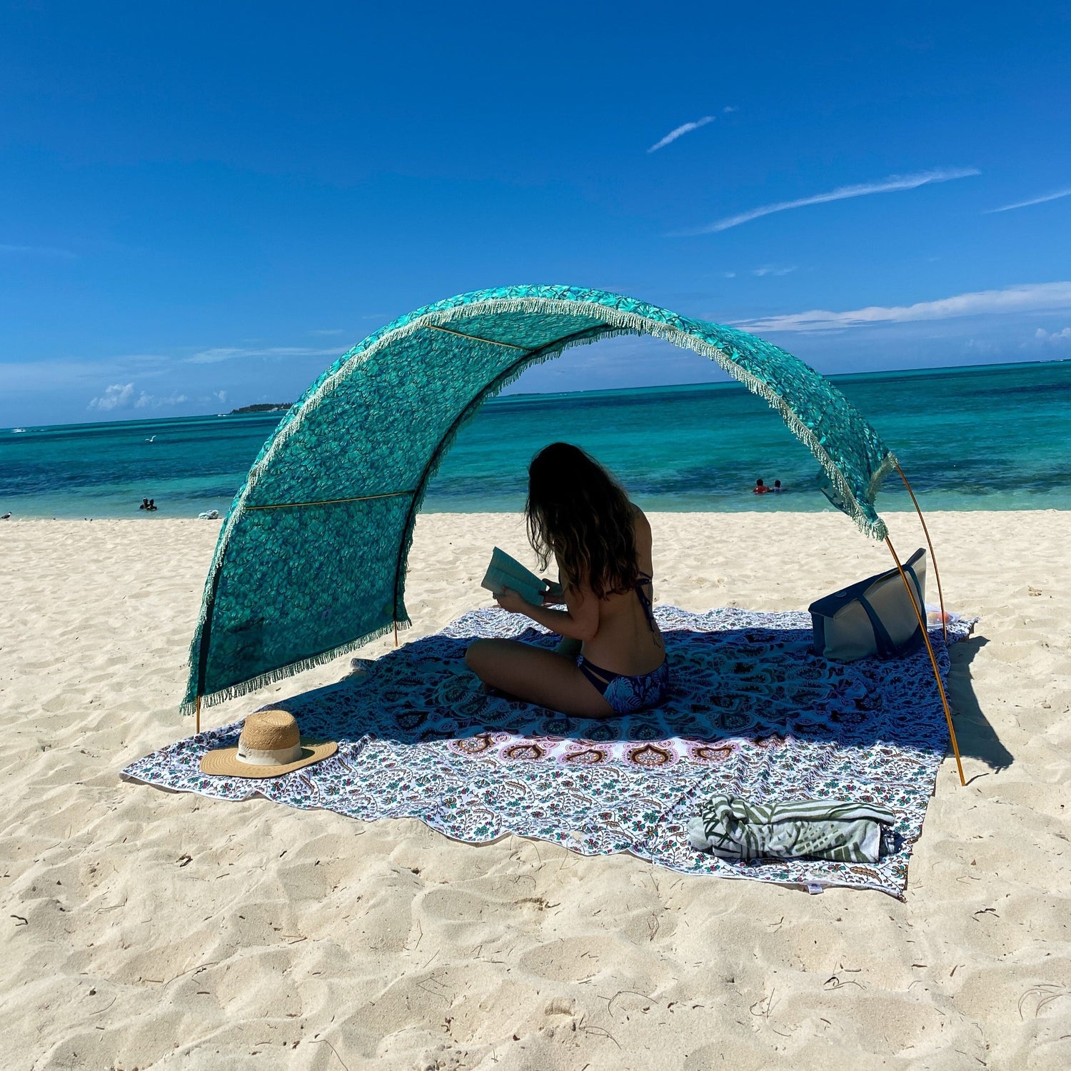 Woman reading under a Tortoise Shell Suniela cabana on a sandy beach, showing full shade coverage and adjustable canopy by the ocean.