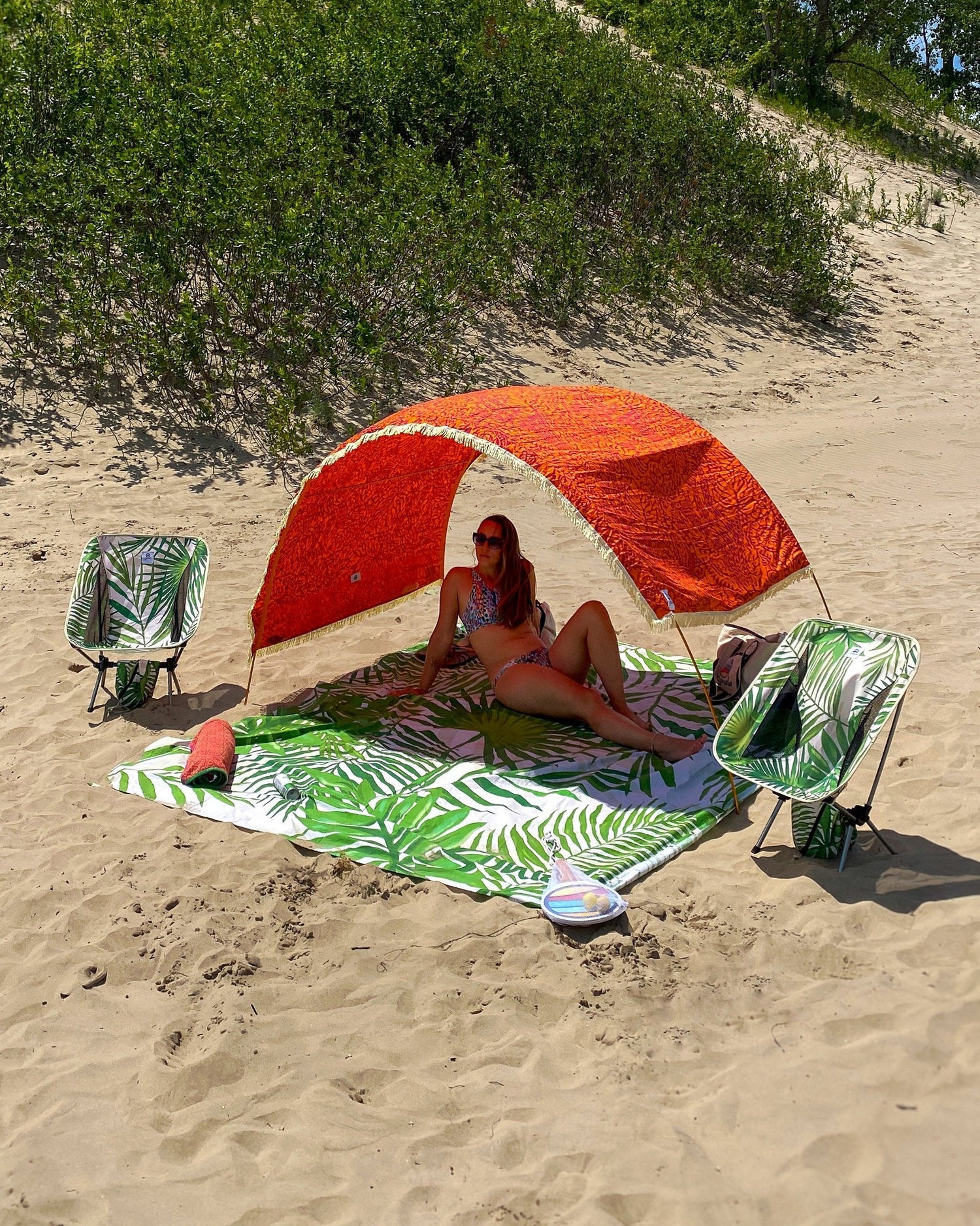 Tribal Jungle Suniela beach cabana set up on a sandy beach with a woman relaxing underneath, paired with packable beach chairs and a matching beach mat