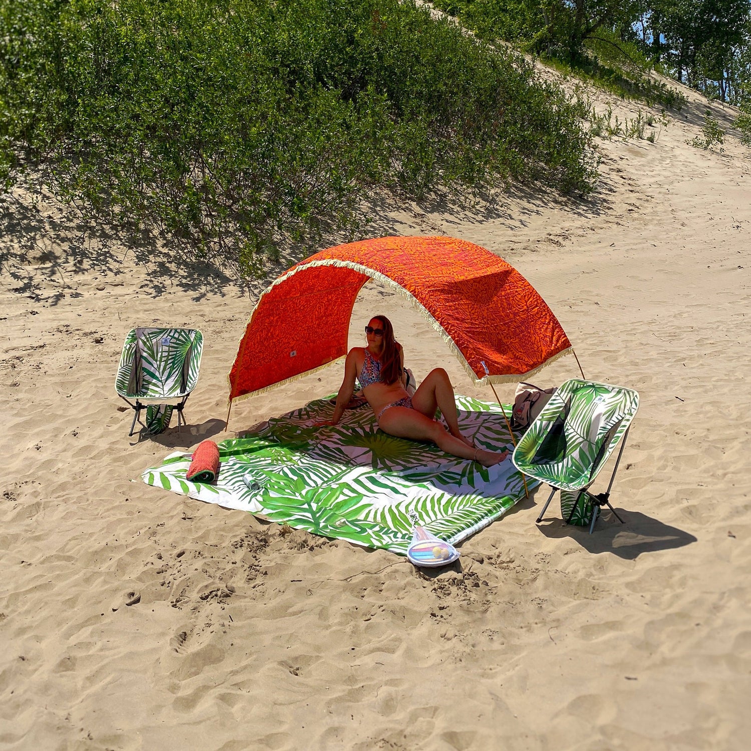 Tribal Jungle Suniela beach cabana set up on a sandy beach with a woman relaxing underneath, paired with packable beach chairs and a matching beach mat