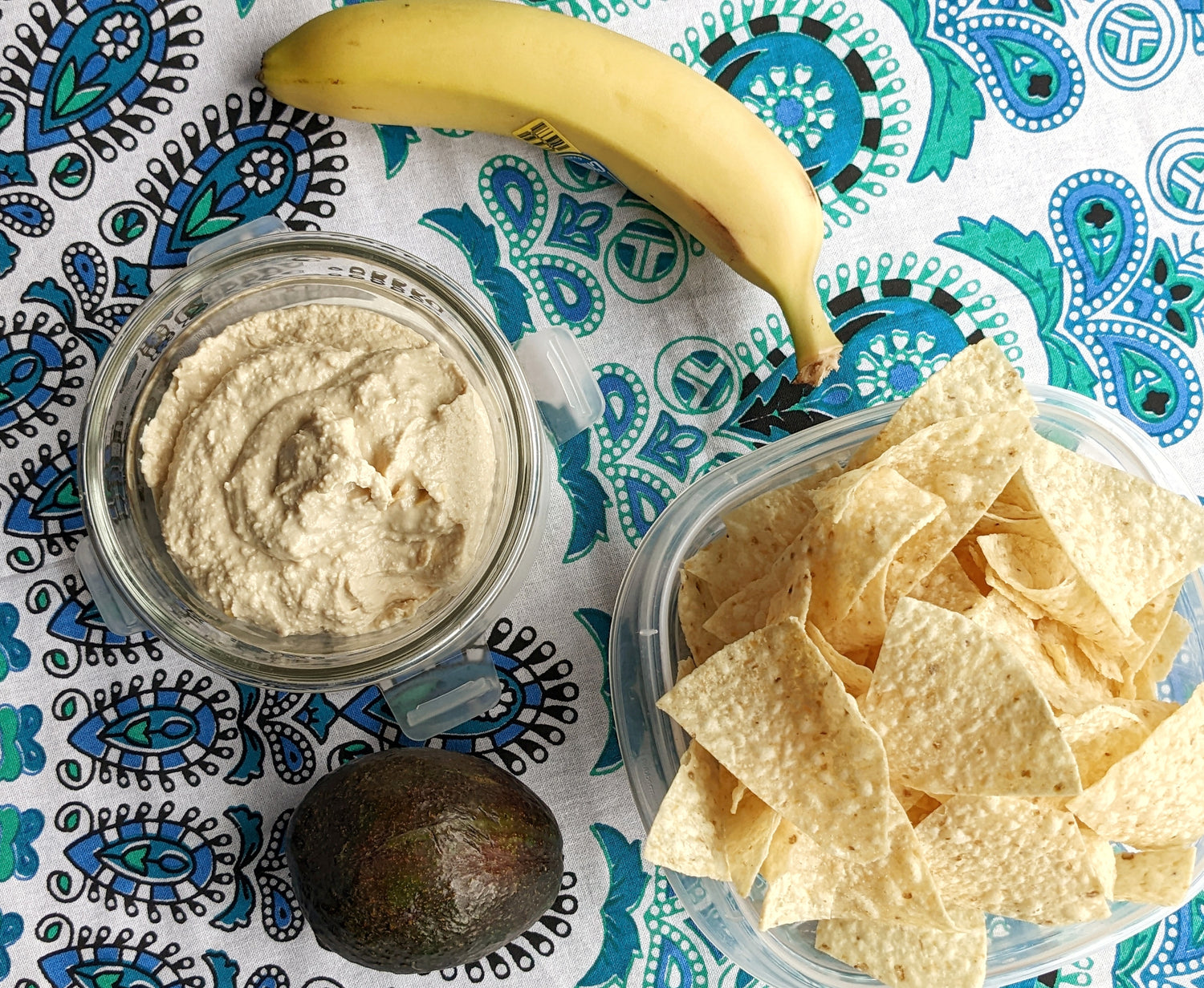 A healthy beach snack: bowl of dip with tortilla chips and a banada on a blue mandala beach mat