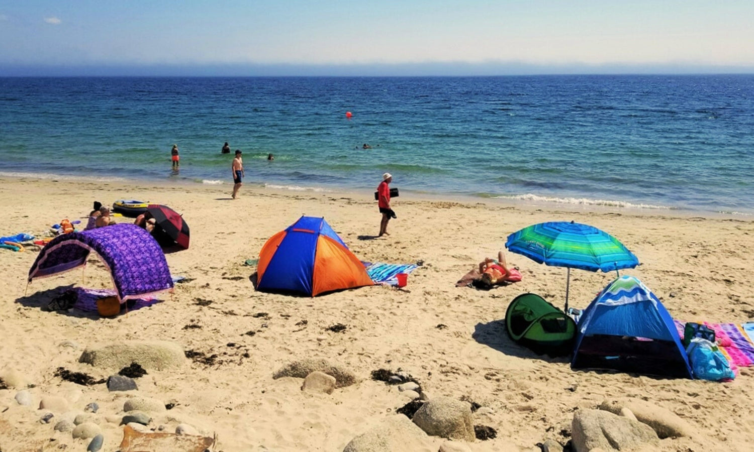 A beach with a synthetic beach tent, sun parasol, and arched Suniela shade cabana, turquoise water in the background and a few people swimming