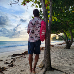 Man stands on a tropical beach carrying a pink suniela shade in it's compact carry bag over his shoulder