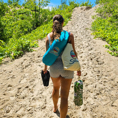 Woman walking up a sandy dune carrying a turquoise Suniela beach cabana over her shoulder, holding sandals and a packable beach chair, highlighting lightweight portability.