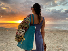 A woman stands on a sandy beach at sunset facing the ocean, carrying a Suniela shade cabana bag in a hand dyed colourful pattern