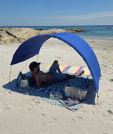 A man relaxes under a blue Suniela beach canopy on a cotton mat on a sandy beach with the ocean and rocks in the background.