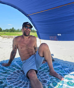A man sitting shirtless on a mandala beach mat under a blue beach tent on a sandy beach.