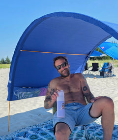 A man wearing sunglasses sits under a blue beach canopy on a sandy beach, holding a water bottle. The canopy provides shade, with other beach chairs and umbrellas visible in the background under a clear blue sky.