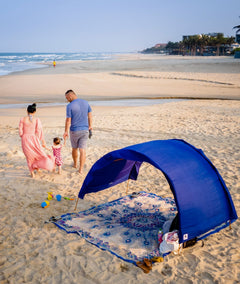 A family of three walks on a sandy beach while a blue beach tent and beach mat are set up in the foreground.