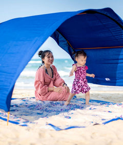 A woman and a young child sitting under a bright blue beach canopy on a sandy beach with the ocean in the background.