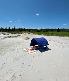 A person relaxes under a blue beach cabana on a sandy beach with green dunes and a blue sky in the background.