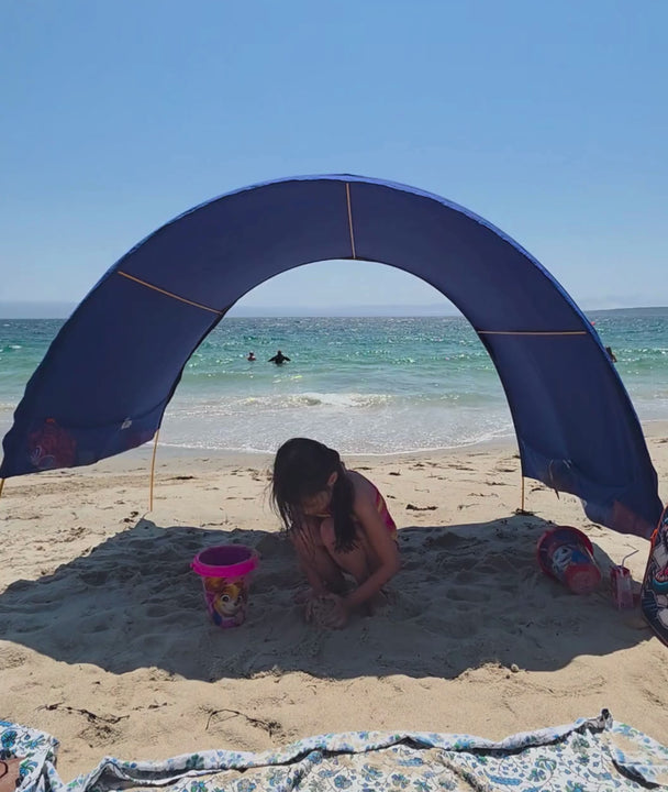 a little girl makes sandcastles in the shade of a blue arched Suniela shade cabana while it blows gently in the wind