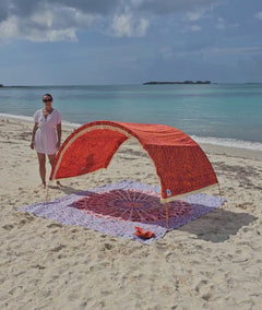 a woman demonstrates the adjustable shade canopy on an arched Suniela Beach cabana, on a sandy beach with turquoise sea in the background