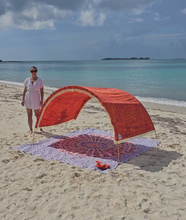 a woman demonstrates the adjustable shade canopy on an arched Suniela Beach cabana, on a sandy beach with turquoise sea in the background