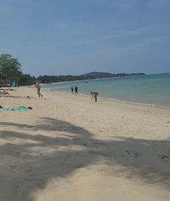a panoramic view of a sandy beach in Thailand, with people swimming in turquoise water. The video pans to a  turquoise shade cabana with a man napping beneath, with the pockets filled with personal items.