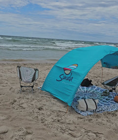 a sandy beach with lots of waves and a Suniela shade cabana moving a lot in the wind but not blowing away.  A man sits on a palm beach chair beneath