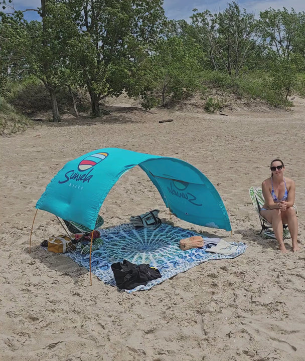 a woman sits on a palm camping chair next to a Suniela shade cabana that's withstanding extremely windy weather on a Canadian beach.