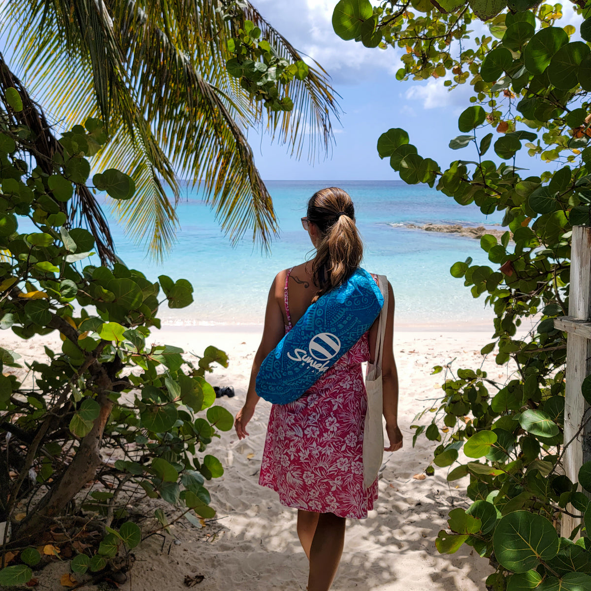 woman carrying a compact Suniela shade cabana in it's batik cotton carry bag, over her shoulder. She wears a pink dress and is walking through tropical foliage toward turquoise water.
